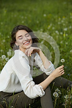 Portrait of a girl sitting in a field on the spring grass among dandelion flowers. Cheerful girl enjoys Sunny spring weather.