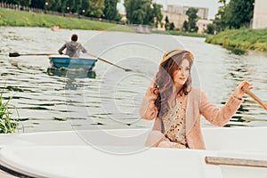 portrait girl is sitting in a white boat on the river. in the distance one sees a retiring boat with a rower