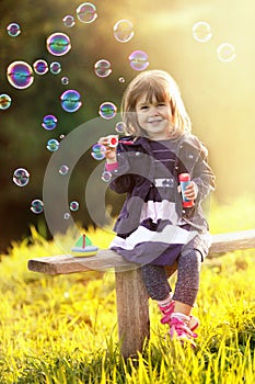 Portrait of a girl sitting on a wooden bench blows bubbles in th