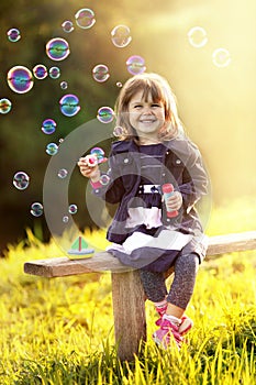 Portrait of a girl sitting on a wooden bench blows bubbles in th