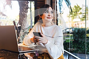 Portrait girl with smartphone in her hands, sitting cafe in front of computer. Woman sitting cafe at table, holding smartphone