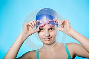 Portrait of a girl sportswoman in a bathing cap and glasses. The girl wears diving goggles. A bright blue background.