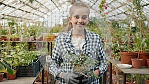 Portrait of girl standing inside greenhouse holding pot plant smiling and looking at camera