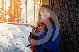 Portrait of a girl in a Sunny winter forest, which stands near a tree in a blue jacket and a red scarf