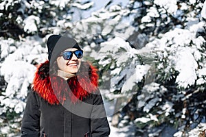 Portrait of a girl teenager wearing black and red coat standing among fir trees covered with snow at the winter park. Young woman