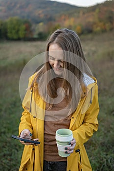 Portrait of girl with thermo cup and smartphone on autumn landscape background. Young woman drinks tea while walking in nature