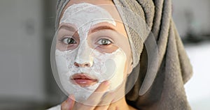 Portrait of girl with towel on hairs applying white moisturising mask on face.