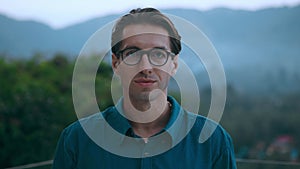 Portrait glad 30 millennial male smiling and adjusting his glasses on backdrop of mountains and beautiful nature in the