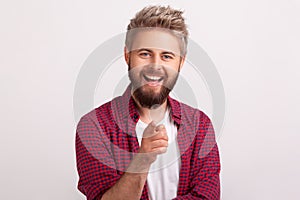 Portrait of glad bearded male in red shirt sincerely smiling at camera and gesturing hey you sign
