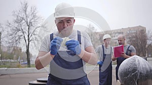 Portrait of glad caucasian man in white helmet and blue uniform listening to money rustle and walking away. Deliveryman