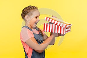 Portrait of glad curious little girl with braid in denim overalls peeking into gift box and laughing from happiness photo