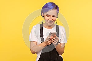 Portrait of glad hipster girl using cell phone, texting or reading message with good news. yellow background, indoor studio shot