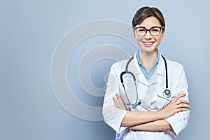 Portrait of glad smiling doctor in white uniform standing with crossed hands on blue clinic background