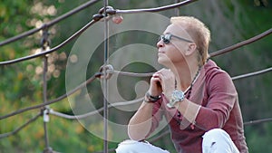 Portrait glamorous handsome blond man in dark glasses sits on suspension bridge over river