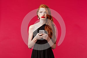 Portrait of gloomy upset gorgeous redhead girl in black dress, sobbing and grimacing unhappy, holding mobile phone