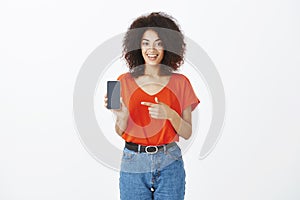 Portrait of good-looking african-american shop assistant in trendy red t-shirt and jeans, showing smartphone and