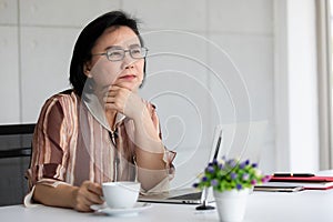 Portrait of good looking and kindly warm feeling Asian senior woman wearing eyeglasses sitting and drinking coffee at working desk
