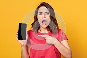 Portrait of good looking young brunette female showing smartphone screen and points at it with her finger, woman has schocked