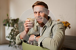 Portrait of good-looking young man with cup of coffee, sitting on chair in cafe, smiling and relaxing with his