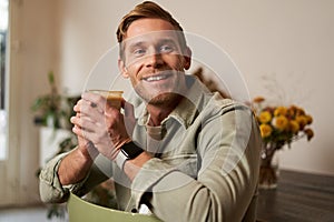 Portrait of good-looking young man with cup of coffee, sitting on chair in cafe, smiling and relaxing with his