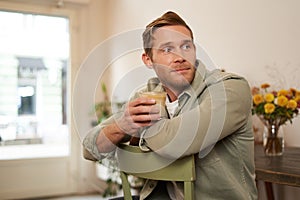 Portrait of good-looking young man with cup of coffee, sitting on chair in cafe, smiling and relaxing with his