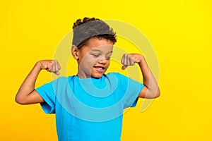 Portrait of good mood strong schoolboy with afro hair wear blue t-shirt look at biceps clenching fists isolated on vivid