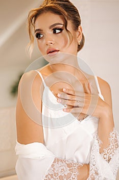 Portrait of gorgeous bride in white silk robe. Woman with beautiful make up and hairstyle. Morning of bride. Wedding