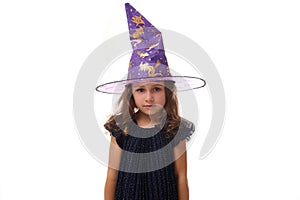 Portrait of gorgeous confident little girl wearing a wizard hat and dressed in stylish carnival dress, looking at camera posing