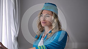 Portrait of gorgeous confident woman in stewardess uniform looking out window opening curtain and smiling. Happy