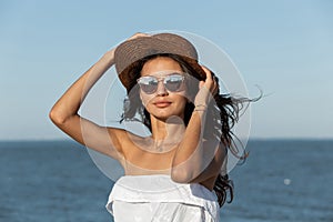 Portrait of gorgeous dark-haired girl in white dress, sunglasses and hat near the sea on a sunny day.