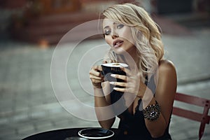 Portrait of gorgeous elegant blond woman sitting at the table in the nice street cafe holding a cup with frothy latte