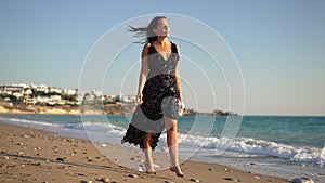 Portrait of gorgeous graceful barefoot woman in elegant dress walking in slow motion on sandy Mediterranean sea beach