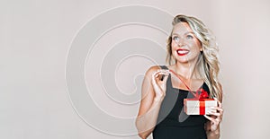 Portrait of gorgeous happy blonde young woman with bright makeup in evening dress with gift box in hands on beige background