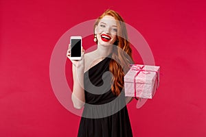 Portrait of gorgeous redhead woman in stylish black dress, laughing and smiling, red lipstick, showing mobile phone