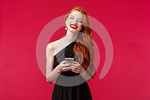 Portrait of gorgeous smiling woman with ginger hair in elegant black prom dress, holding smartphone, messaging, using