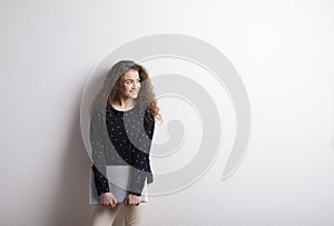 Portrait of a gorgeous teenage girl with curly hair, holding notebook, leaning against wall. Studio shot, white