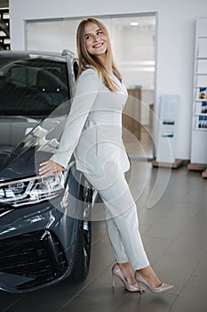 Portrait of gorgeous woman in showroom. Beautiful salesperson standing by car. Blond hair female after buying car