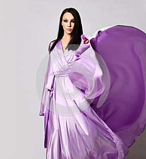 Portrait of graceful brunette woman in lilac evening maxi dress silk gown stands with the hem fluttering in the wind