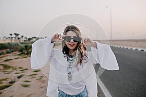 Portrait of graceful stylish girl in black sunglasses and white blouse having fun next to highway in summer. Charming