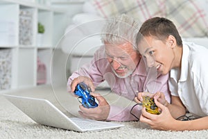 Portrait of grandfather and grandson lying on floor and playing computer games on laptop