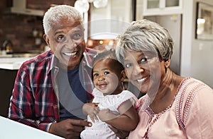 Portrait Of Grandparents Sitting With Baby Granddaughter Around Table At Home