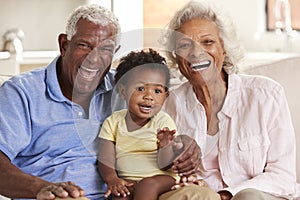 Portrait Of Grandparents Sitting On Sofa At Home Playing With Baby Granddaughter