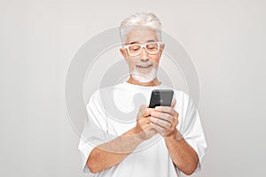 Portrait of gray-haired man in white T-shirt holding mobile phone in hand with happy smiling face. Person with smartphone