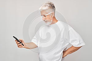 Portrait of gray-haired man in white T-shirt looks on mobile phone and thinks. Person with smartphone