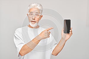 Portrait of gray-haired man in white T-shirt showing blank mobile phone screen with excited face. Person with smartphone