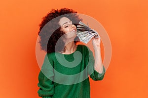 Portrait of greedy woman with Afro hairstyle wearing green casual style sweater holding and smelling fan of hundred dollar bills,