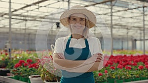 Portrait of the greenhouse gardener looking at the camera wearing hat. Female worker crossing her arms standing on the