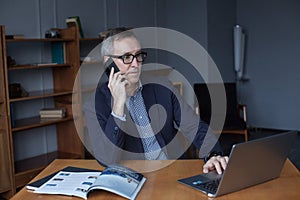 Portrait of grey hair elderly man sitting front table with laptop, talking on mobile phone