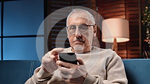 Portrait of grey-haired aged man using mobile app in smartphone in home, resting in weekend evening
