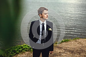 Portrait of the groom in nature in the summer. A handsome man in a suit stands on the shore of the pond and looks into the